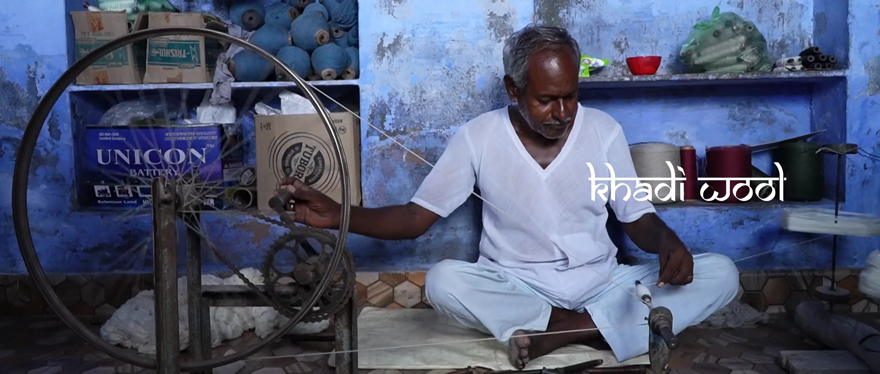 An artisan sitting cross-legged spinning khadi wool on a charkha in a blue-walled room